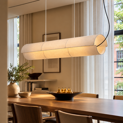 A modern cylindrical pendant light with a warm white fabric-covered shade hangs over a wooden dining table in a softly lit dining room.