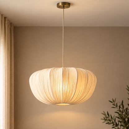 A modern pendant light with a ribbed, pleated shade in a warm cream or off-white color, suspended by a slim rod from a brass ceiling canopy. The lamp is shown illuminated in a warm, cozy room setting with a beige wall, a curtain on the left, and a plant on the right.